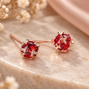 Puede incluir: Un par de pendientes de botón en oro rosa con gemas redondas y facetadas de color rojo intenso. Cada piedra está asegurada por un engaste metálico en forma de jaula con barras finas y entrecruzadas que envuelven la gema, con un poste recto.