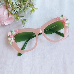 Può includere: Occhiali rosa a occhi di gatto con lenti verdi, adornati con piccoli fiori rosa e bianchi e perle sulla montatura superiore. Gli occhiali hanno uno stile retrò e sono su uno sfondo bianco con accenti floreali.
