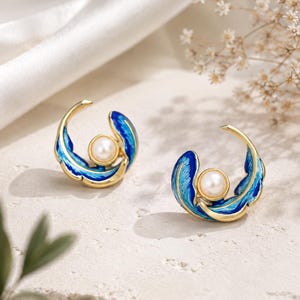 Può includere: Un paio di orecchini dorati con un design a mezzaluna, dettagli in smalto blu e una perla bianca centrale. Gli orecchini presentano una struttura curva simile a una piuma che avvolge la perla, creando una silhouette circolare.