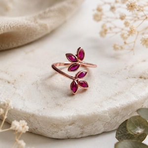 May include: A rose gold bypass ring featuring four marquise-cut deep pink stones arranged in two pairs to resemble leaves. The open-ended band design allows the two ends to overlap slightly, showcasing the vibrant pink stones at each terminal point.