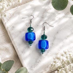 Peut inclure: Une paire de boucles d'oreilles pendantes composée d'une perle carrée en verre bleu au centre, encadrée par des perles en cristal vert foncé à facettes. Les boucles sont fixées à des crochets en métal argenté, formant un bijou aux couleurs vives.