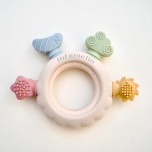 May include: A baby teether toy featuring a light pink wooden ring and colourful silicone attachments. The attachments include a blue airplane, a green car, a pink star, and a yellow sun. The word "Infantella" is printed on the ring.