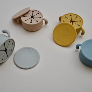 May include: Four silicone snack bowls in beige, yellow, and blue, each with a matching lid. The lids have a starburst design and the bowls have handles. The bowls are designed for snacks.
