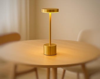 May include: A gold-colored, modern table lamp with a cylindrical base, a slender stem, and a circular top. The lamp is illuminated, casting a warm, inviting glow. The lamp is sitting on a wooden table.