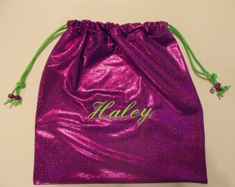 May include: A magenta drawstring bag with the name "Haley" embroidered in green script. The bag features a green drawstring with decorative beads and a knot. The bag is made of a shiny, textured material. The bag is a vibrant magenta colour.