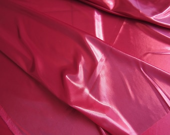 May include: Close-up of a vibrant, shiny, fuchsia-coloured fabric. The material is draped, creating soft folds and highlights. The fabric has a subtle texture and a glossy finish, reflecting light.