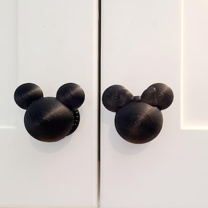 May include: Two black cabinet knobs in the shape of Mickey Mouse's head. The knobs are mounted on white cabinet doors. The knobs have a textured surface and a classic, cartoonish design.