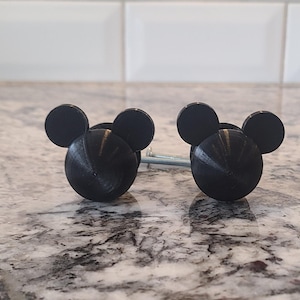 May include: Two black cabinet knobs shaped like a mouse head with circular ears. The knobs are made of a solid material and have a textured surface. The knobs are shown on a marble countertop.