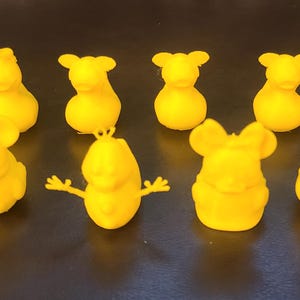 May include: A collection of nine yellow figurines, each with a unique design. The figurines include mouse-like characters with large ears, and other abstract shapes. The objects are arranged on a dark surface.