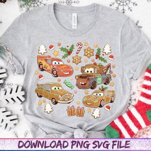 May include: A light gray t-shirt featuring a colorful graphic of four cartoon cars designed as gingerbread cookies, with Christmas-themed decorations. The design includes candy canes, holly, and gingerbread house cookies. The shirt has a crew neck and short sleeves.