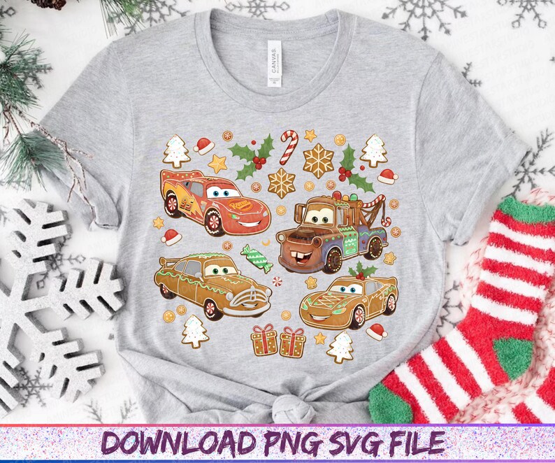 May include: A light gray t-shirt featuring a colorful graphic of four cartoon cars designed as gingerbread cookies, with Christmas-themed decorations. The design includes candy canes, holly, and gingerbread house cookies. The shirt has a crew neck and short sleeves.