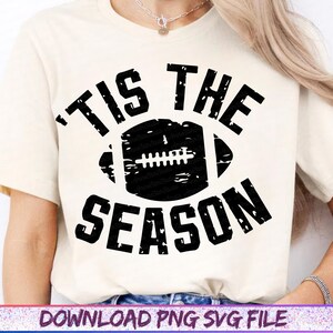 May include: Cream-colored t-shirt with a distressed black football graphic and the words "TIS THE SEASON". The design is centered on the shirt, perfect for football fans.