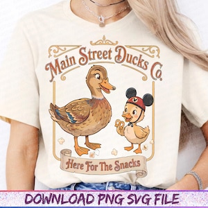 May include: Cream-colored t-shirt featuring a vintage-style graphic. The design includes two cartoon ducks, one wearing a Mickey Mouse hat, and the text "Main Street Ducks Co." and "Here For The Snacks" in a decorative frame.