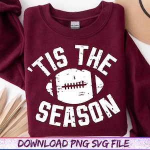 May include: A maroon sweatshirt with the text "'TIS THE SEASON" in white, featuring a white football graphic. The sweatshirt is a Gildan brand and is folded. The bottom of the image has the text "DOWNLOAD PNG SVG FILE".