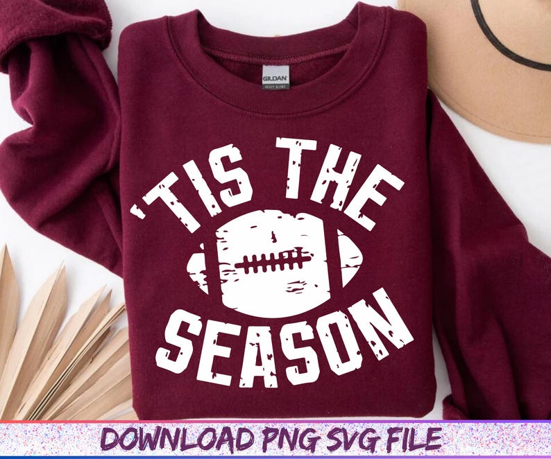 May include: A maroon sweatshirt with the text "'TIS THE SEASON" in white, featuring a white football graphic. The sweatshirt is a Gildan brand and is folded. The bottom of the image has the text "DOWNLOAD PNG SVG FILE".