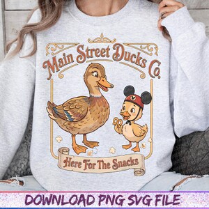 May include: A light gray sweatshirt featuring a cartoon duck and duckling with the text "Main Street Ducks Co." and "Here For The Snacks." The duckling wears a Mickey Mouse hat and holds a pretzel.