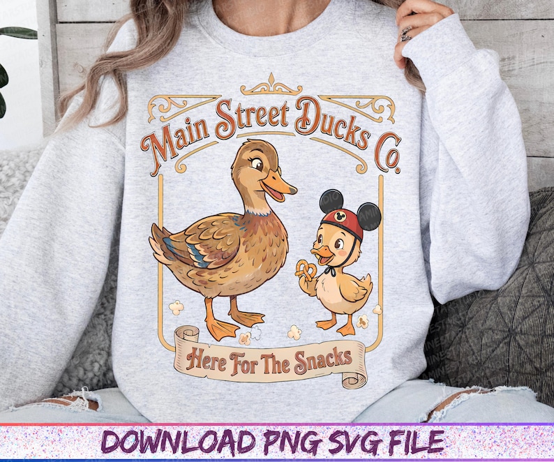 May include: A light gray sweatshirt featuring a cartoon duck and duckling with the text "Main Street Ducks Co." and "Here For The Snacks." The duckling wears a Mickey Mouse hat and holds a pretzel.