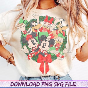 Op de afbeelding: Crèmekleurig T-shirt met een feestelijk kransontwerp met Mickey Mouse, Minnie Mouse, Donald Duck, Daisy Duck en Goofy in kerstoutfits. De krans is versierd met rode strikken en zuurstokstrepen. De tekst "DOWNLOAD PNG SVG FILE" staat onderaan.