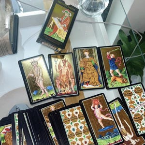 May include: A deck of Visconti Tarots cards with gold accents and illustrations, alongside the black and gold box. The cards are spread out on a reflective surface, showcasing various detailed images.
