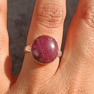 May include: A silver ring with a large, round, deep red gemstone. The ring is on a hand with fair skin, the fingers slightly curved. The gemstone has a polished, smooth surface. The ring is a simple, elegant design.