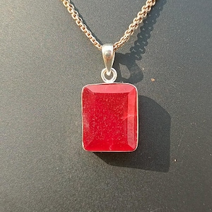 May include: A silver necklace featuring a rectangular, bright red gemstone pendant. The pendant is set in a silver bezel and suspended from a fine chain. The necklace is displayed on a black mannequin.