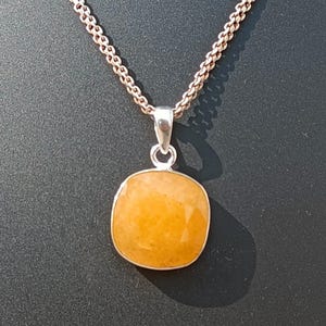 May include: A silver chain necklace featuring a square, orange gemstone pendant. The pendant is set in a silver bezel and hangs from a delicate chain. The necklace is displayed on a black mannequin.