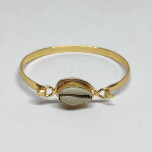 Puede incluir: Una pulsera rígida de metal dorado con una concha de cauri blanca en el centro. La concha está montada en un engaste de metal dorado que se conecta a la banda circular fina y lisa mediante pequeñas anillas metálicas a ambos lados.