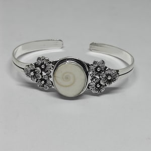 May include: A silver-toned cuff bracelet featuring a central oval-shaped white shiva eye shell. The metal band is decorated on both sides of the center stone with detailed floral motifs, showcasing a polished finish and an open-ended design.