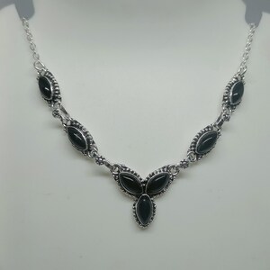 Puede incluir: Un collar de tono plateado que presenta una serie de piedras negras con corte marquesa engastadas en marcos de metal ornamentados. El diseño incluye cinco engastes individuales conectados por pequeños eslabones circulares.