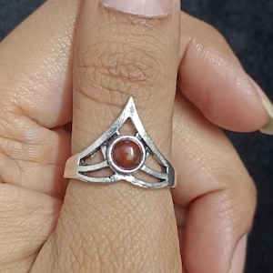 May include: A silver ring with a triangular top and a circular, reddish-brown stone set in the center. The ring is worn on a thumb against a dark background.
