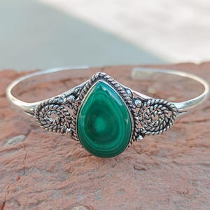 May include: A silver bracelet with a striking, teardrop-shaped malachite stone in vivid green. The stone is set in an intricate silver setting with swirling patterns. The bracelet has a simple, open design, suitable for various wrist sizes.