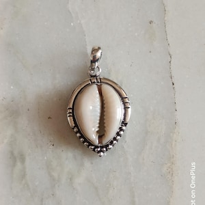 May include: A silver pendant with a cowrie shell. The shell is white and brown, set in a decorative silver frame with a beaded edge. The pendant has a loop for a chain and is shown on a light surface.