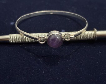 May include: A silver bangle bracelet with a round, polished, purple amethyst gemstone set in the center. The bracelet is resting on a silver tool with a textured handle. The background is a dark blue.