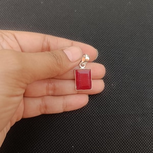 Puede incluir: Un colgante rectangular con una vibrante gema roja engastada en un marco de metal plateado. La gema tiene una superficie lisa y pulida. El colgante está suspendido de un pequeño lazo en la parte superior.