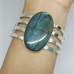 Puede incluir: Brazalete de plata con una gran piedra ovalada de labradorita con tonos iridiscentes azules y verdes. El brazalete tiene un diseño multibanda, con cuatro bandas de plata paralelas.