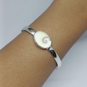 Könnte beinhalten: Ein silbernes Armreif-Armband mit einem ovalen, cremefarbenen Mittelstück. Das Mittelstück weist ein Spiralmuster auf. Das Armband wird am Arm einer Person getragen.