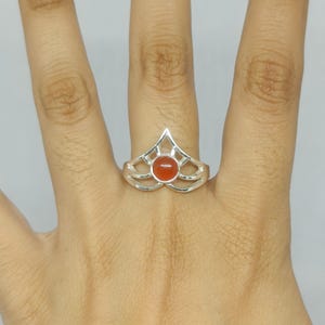 May include: A silver ring featuring a central round orange stone set within a geometric, openwork frame that rises to a point. The band is constructed from polished metal, showcasing a symmetrical design that encircles the finger with open spaces.