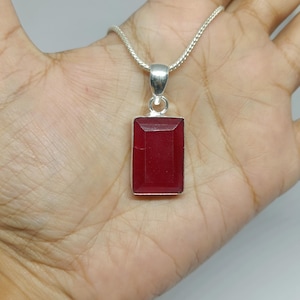 May include: A rectangular, deep red gemstone pendant set in a polished silver frame, suspended from a matching silver chain. The stone features a faceted surface that catches the light, and the pendant is attached to the chain by a silver bail.