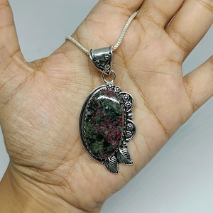 Puede incluir: Un colgante de forma ovalada con una piedra moteada en verde y rosa oscuro, engastada en un marco de plata decorativo con motivos de hojas y flores. El colgante cuelga de una fina cadena de plata con intrincados detalles metálicos.