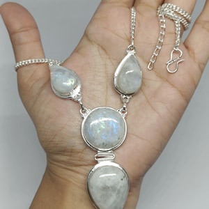 Puede incluir: Un collar de plata con cuatro cabujones de piedra lunar engastados en metal pulido. El diseño incluye dos piedras en forma de pera en la parte superior, una gran piedra circular central y una piedra en forma de pera en la parte inferior, unidas por una cadena.