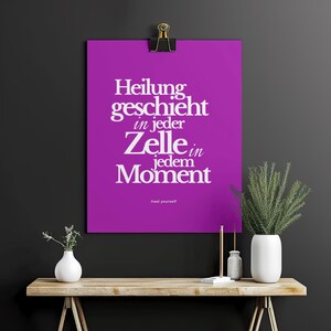 May include: A purple poster with the German text "Heilung geschieht in jeder Zelle in jedem Moment" in white, held by a black clip. The phrase "heal yourself" is at the bottom. A wooden table holds vases and plants.