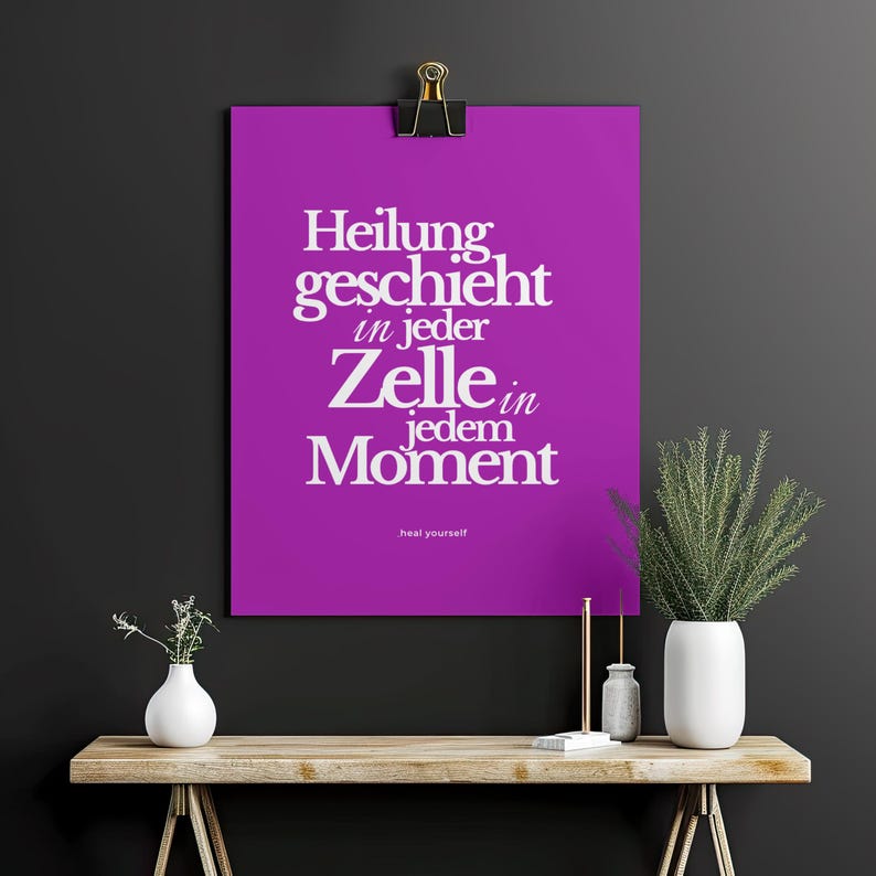 May include: A purple poster with the German text "Heilung geschieht in jeder Zelle in jedem Moment" in white, held by a black clip. The phrase "heal yourself" is at the bottom. A wooden table holds vases and plants.