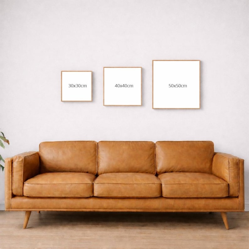 Op de afbeelding: Drie ingelijste vierkante kunstdrukken met witte achtergronden en gouden frames, van 30x30cm, 40x40cm en 50x50cm, worden boven een bruine leren bank weergegeven. De bank heeft drie kussens en houten poten.