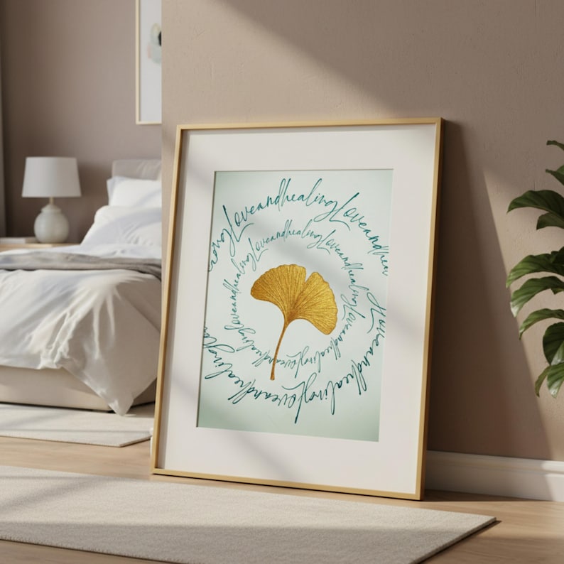 Op de afbeelding: Een ingelijste kunstafdruk met een gouden ginkgoblad-illustratie. Het blad is omgeven door turquoise tekst met de tekst "love and healing". De gouden lijst vormt een aanvulling op het kunstwerk, dat op een lichtgekleurde muur wordt getoond.
