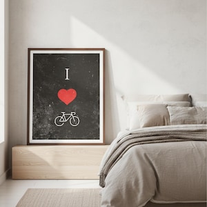 Op de afbeelding: Ingelijste kunstdruk met een zwarte achtergrond, met een rood hart en een witte fietsafbeelding, met de woorden "I" boven het hart. De print zit in een houten lijst en wordt in een slaapkamer getoond.