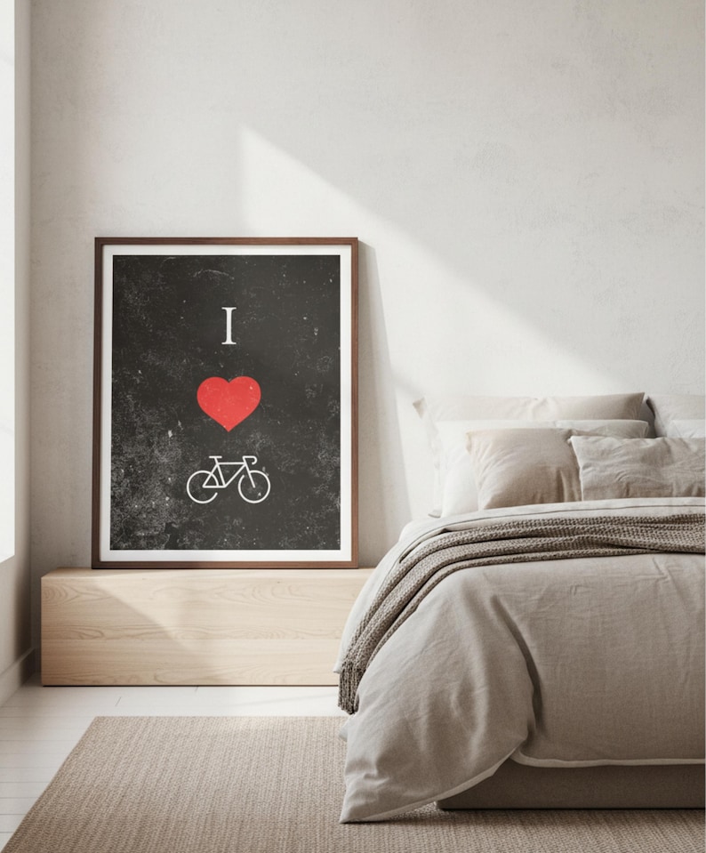 Op de afbeelding: Ingelijste kunstdruk met een zwarte achtergrond, met een rood hart en een witte fietsafbeelding, met de woorden "I" boven het hart. De print zit in een houten lijst en wordt in een slaapkamer getoond.