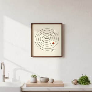 Op de afbeelding: Ingelijste print met een cr&egrave;mekleurige achtergrond. Het kunstwerk heeft een zwart spiraalontwerp met de woorden "me" en "you" en een klein rood hart. De lijst is donkerbruin.