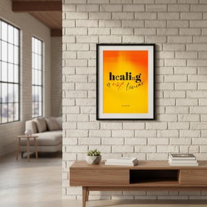 May include: A framed art print with the text "healing is not linear" in black against a yellow and orange gradient background. The artwork is displayed on a white brick wall above a wooden console table. A small plant and books are on the table.