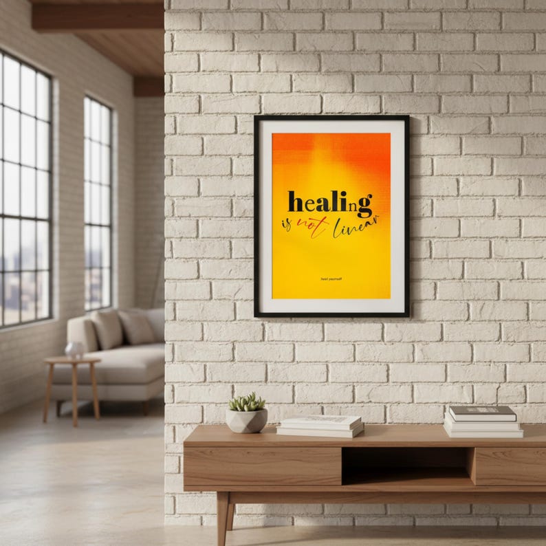 May include: A framed art print with the text "healing is not linear" in black against a yellow and orange gradient background. The artwork is displayed on a white brick wall above a wooden console table. A small plant and books are on the table.