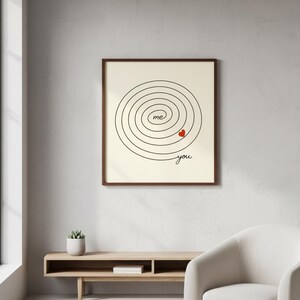 Op de afbeelding: Een ingelijste print met een cr&egrave;mekleurige achtergrond toont een zwart spiraalontwerp. De woorden "me" en "you" zijn in zwarte letters geschreven, met een klein rood hart. De lijst is donkerbruin.